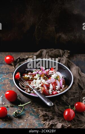 Bol noir avec différentes tomates en tranches, oignon rouge, sense balsamique, salade de thym et de feta, servi avec une fourchette et de la tomate cerise sur une sanche humide Banque D'Images
