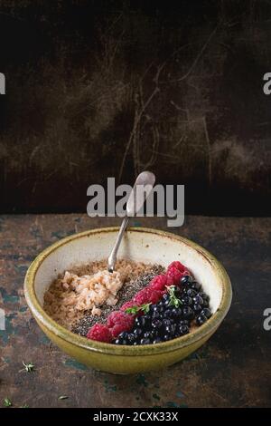 Bol de porridge à flocons d'avoine avec bleuets, framboises et graines de chia, servi avec une cuillère vintage sur fond texturé de bois ancien. Petit déjeuner rustique t Banque D'Images