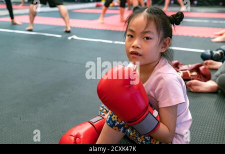 7 ans enfant fille apprenant la boxe thaï ou Muay Thai dans la salle de gym. Assis sur le sol, portant des gants de boxe et regardant la caméra. Banque D'Images