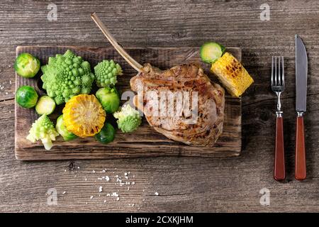 Steak de veau grillé tomahawk avec légumes choux de Bruxelles, romanesco et épis de maïs servi avec couverts en bois servant de planche à découper de vieux wo Banque D'Images