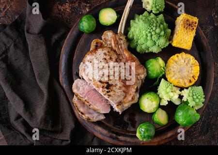 Steak de tomahawk de veau grillé en tranches avec légumes choux de bruxelles, romanesco et épis de maïs servis sur une planche à découper en bois sur un vieux bac en bois Banque D'Images