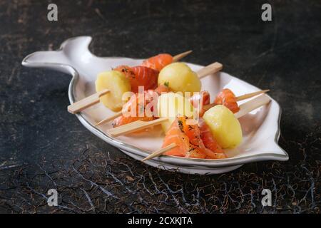 Délicieux hors-d'œuvre avec saumon fumé salé et pommes de terre cuites sur brochettes servies sur une assiette en forme de poisson blanc sur fond de texture sombre. Banque D'Images
