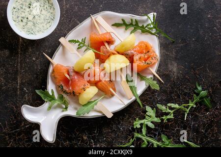 Apéritif délicieux saumon salé et fumé avec des pommes de terre en brochettes servi avec sauce crémeuse et roquette sur forme de poisson blanc sur plaque Banque D'Images