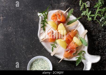 Apéritif délicieux saumon salé et fumé avec des pommes de terre en brochettes servi avec sauce crémeuse et roquette sur forme de poisson blanc sur plaque Banque D'Images