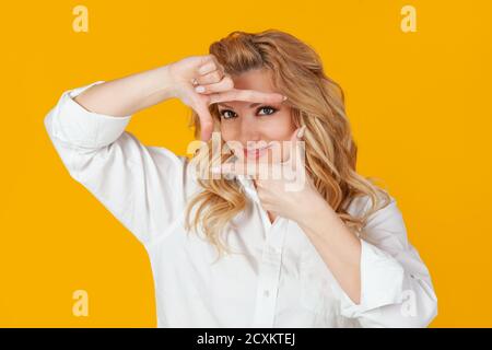 Une blonde en chemise blanche, une femme européenne d'âge moyen, un concepteur créant un nouveau projet, regarde les images et sourit, et se tient sur un fond jaune pour créer la photo parfaite Banque D'Images