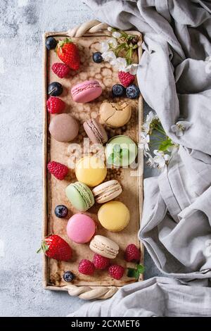 Variété de macarons de desserts doux français colorés avec différentes garnitures servies sur un plateau en terre cuite avec fleurs printanières, baies, ove textile Banque D'Images