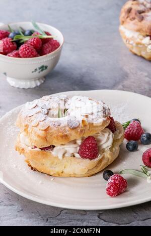 Gâteau de chax maison Paris Brest avec framboises, amande, sucre en poudre, romarin sur plaque blanche avec baies sur fond de texture grise. FR Banque D'Images