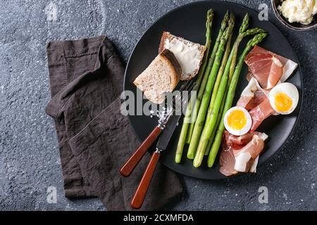 Asperges vertes cuites avec un demi-œuf dur, pain coupé en tranches, beurre et bacon au jambon servi avec du sel de mer et des couverts sur une assiette en céramique noire sur du eston foncé Banque D'Images