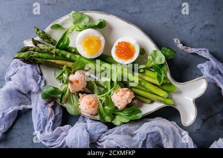 Asperges vertes cuites avec demi-oeuf dur, salade, caviar rouge et saumon pate servi sur plaque de forme de poisson blanc avec de la gaze sur textile bleu gris Banque D'Images