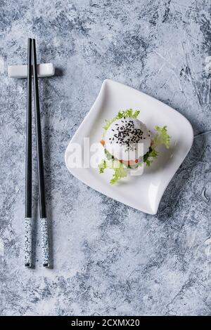 Mini-hamburger à sushi au riz avec saumon fumé, salade verte et sauces, sésame noir servi sur une assiette carrée blanche avec des baguettes sur fond de béton gris Banque D'Images