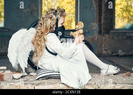 petite ange fille avec des ailes dans un masque respiratoire tenant un ours en peluche dans ses mains. écologie et dévastation concept Banque D'Images