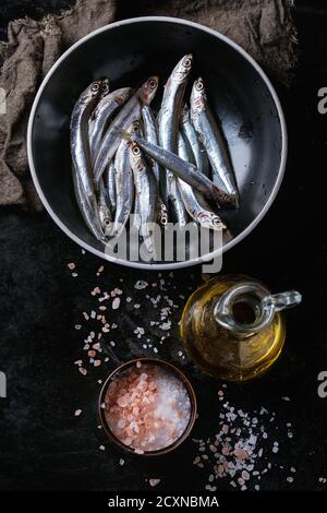 Beaucoup de poissons frais d'anchois crus dans un bol en céramique noire avec du sel rose et de mer et une bouteille d'huile d'olive pour la marinade sur fond de métal noir. Haut v Banque D'Images