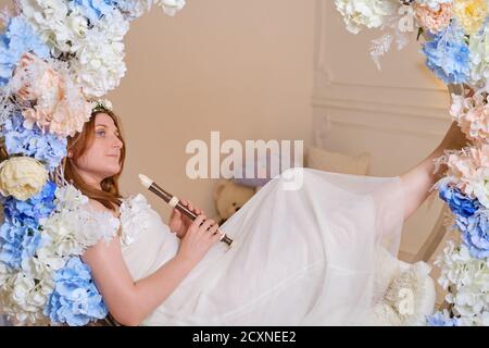 La femme souriante se trouve avec une flûte à bloc dans les fleurs. Studio portrait de la femme heureuse musicienne en robe blanche Banque D'Images