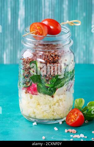 Salade avec quinoa, couscous, épinards, radis, tomates et courgettes dans des pots de maçon en verre, debout avec des légumes frais sur bois turquoise clair b Banque D'Images