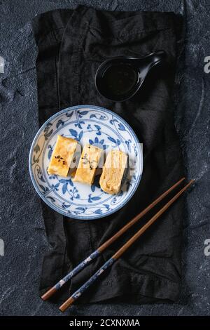 Omelette japonaise Tamagoyaki en tranches de graines d'assaisonnement noires et sauce soja, servie dans une assiette en céramique bleue blanche ornée de baguettes, textile ov Banque D'Images