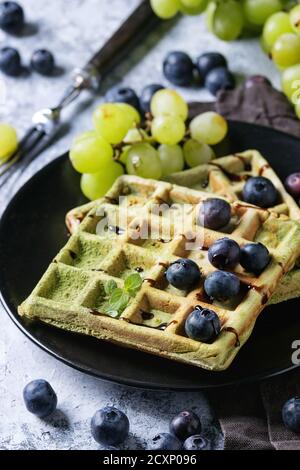 Gaufres belges au matcha vert carré faites maison sur une assiette noire avec raisins mûrs frais, bleuets, menthe, fourchette, serviette textile sur fond de texture gris Banque D'Images