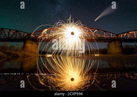 La laine d'acier qui tourne dans un cercle abstrait, des averses d'étincelles jaune vif sur un long pont se reflètent dans l'eau de la rivière sous un ciel étoilé de nuit sombre. Banque D'Images