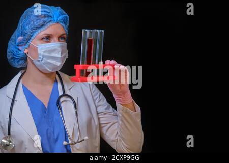 Médecin dans un masque médical avec des tubes de test sanguin pour l'analyse du coronavirus. L'infirmière tient les flacons sur pied dans une main sur un fond sombre, espace de copie. Rés Banque D'Images