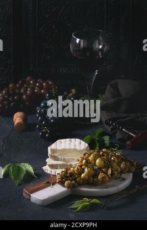 Serving board with sliced camembert cheese and baked bunch of green grapes served with glass of red wine, corkscrew, green leaves over black table. Da Stock Photo