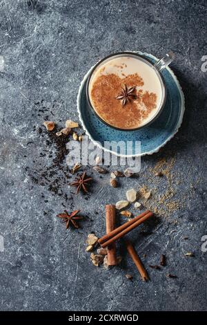 Tasse en verre de thé traditionnel indien masala chai sur soucoupe bleue avec les ingrédients ci-dessus. Cannelle, cardamome, anis, sucre, thé noir sur texture foncée b Banque D'Images