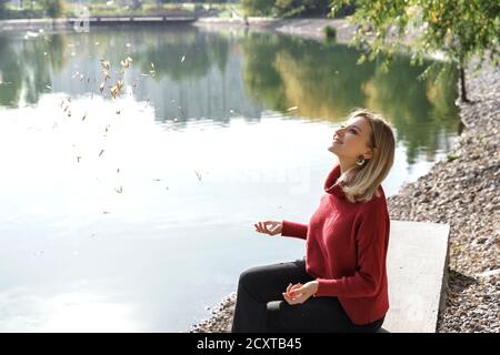 belle jeune femme blonde jette des feuilles vers le haut par le lac et rires. concept d'automne Banque D'Images