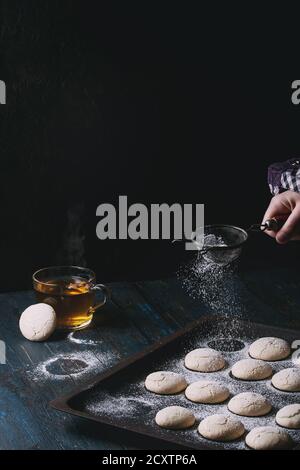 Biscuits faits maison sur le vieux four plateau et tasse de thé chaud sur bleu foncé table en bois. Saupoudrer de sucre en poudre par vintage le tamis dans la main de l'enfant. Dark Banque D'Images