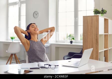 Portrait d'une femme souriante assise au bureau devant un ordinateur portable et s'étirant après le travail. Banque D'Images