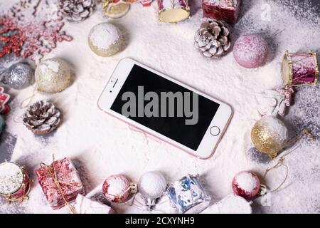 Téléphone blanc avec écran noir à côté des décorations du nouvel an dans la neige. Arbres de Noël, boules, flocons de neige, cadeaux, cônes. Ambiance de Noël. Photo plate. Banque D'Images