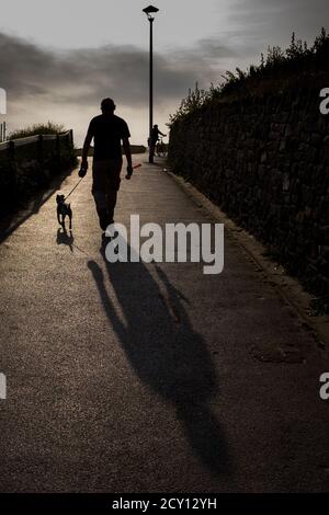 Un homme marche son chien terrier en zigzag alors que le soleil commence à se coucher à l'ouest, au début d'une soirée d'été sur la plage de Southbourne à Bournemouth. Banque D'Images