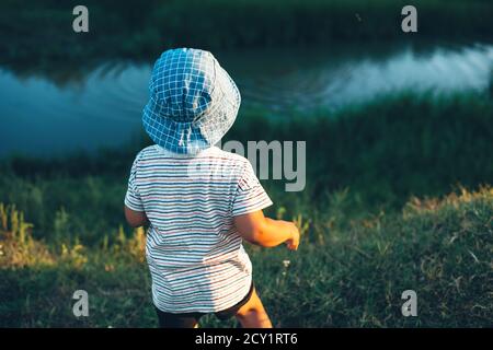 Garçon caucasien regardant une petite rivière et jetant des pierres porter un chapeau bleu sur un terrain vert