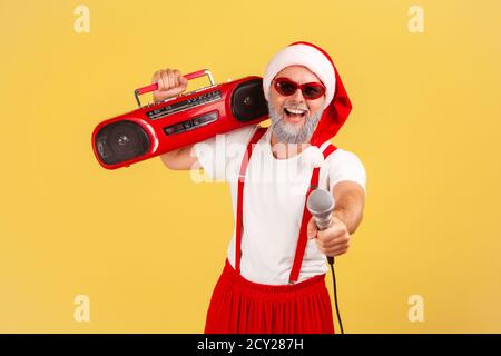 Homme adulte à barbe grise positive dans le chapeau et le pantalon du père noël avec bretelles tenant le magnétophone et tenant le microphone à la caméra. Studio intérieur Banque D'Images