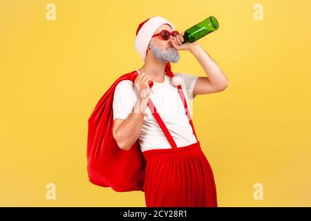 Homme adulte méchant, mal ivre, avec un gros sac rouge et un chapeau de père noël, buvant de la bière à la bouteille, s'amusant, relaxant après les vacances. Studio intérieur Banque D'Images