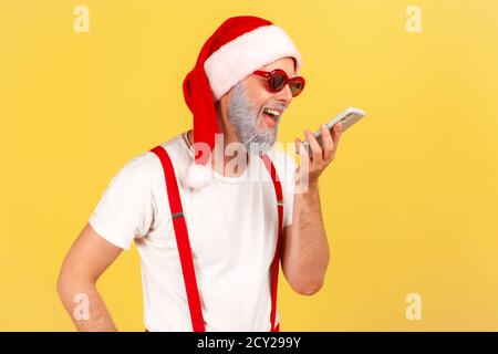 Souriant heureux barbu gris dans le chapeau du père noël et lunettes élégantes tenant le smartphone près de la bouche parlant à l'assistant vocal, laissant le message vocal. Banque D'Images