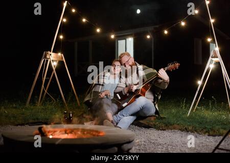 Un jeune couple, une femme caucasienne et un homme barbu aux cheveux rouges, lors d'un pique-nique de nuit avec une guitare et un guimello ont un bon moment Banque D'Images