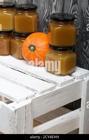 Confiture de fruits en pots. Une citrouille orange se trouve à proximité. Se dresse sur une caisse en bois peinte en blanc. Sur fond de planches de pin noir. Banque D'Images