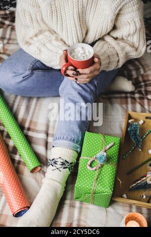 La jeune femme est assise sur un tissu écossais dans un confortable pull blanc en laine tricoté et tient une tasse de cacao avec des guimauves. Nouvel an hygge, préparation pour les vacances. CAN Banque D'Images