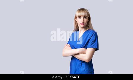 Une jeune femme confiante médecin debout bras croisés. Interne dans des vêtements médicaux bleus posant sur fond gris. Banque D'Images