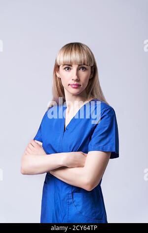 Une jeune femme confiante médecin debout bras croisés. Interne dans des vêtements médicaux bleus posant sur fond gris. Banque D'Images