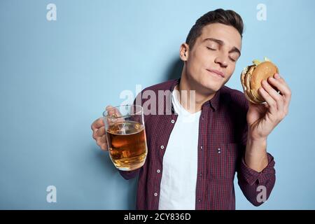 homme gai ivre avec une tasse de bière et un hamburger à la main régime alimentaire style de vie fond bleu Banque D'Images