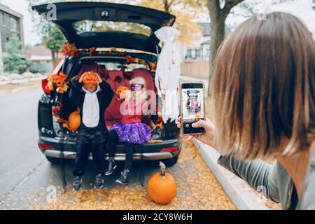 Trick ou tronc. Enfants garçon et fille avec des citrouilles rouges célébrant les fêtes d'Halloween d'octobre dans le coffre de voiture en plein air. Mère prenant des photos d'enfants Banque D'Images