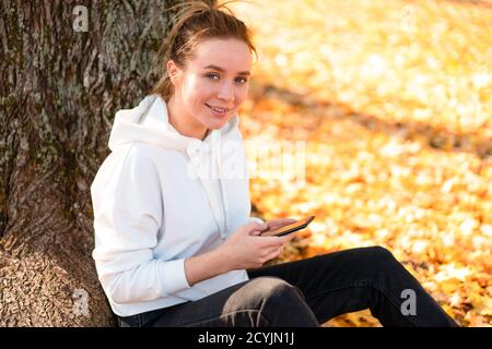 Femme dans un pull à capuche blanc avec une capuche est assis sur le sol dans le parc et la tenue d'un téléphone cellulaire dans les mains. cute girl utilisation smartphone pour communicatio Banque D'Images