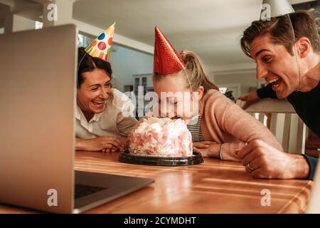 Fille coupant son gâteau d'anniversaire avec sa bouche avec ses parents souriant par. Famille célébrant la fille à la maison pendant la quarantaine Covid-19. Banque D'Images