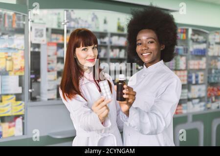 Équipe de deux chimistes multiethniques souriants en pharmacie debout devant des étagères avec des médicaments en pharmacie moderne, tenant une plaquette thermoformée avec des pilules Banque D'Images