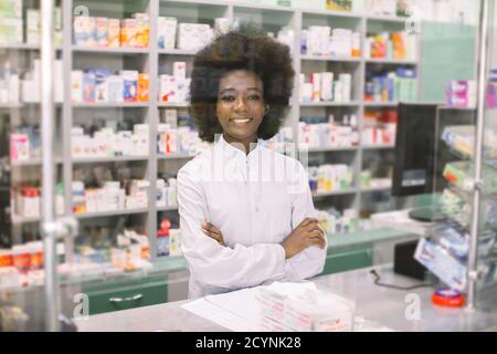 Heureuse jeune femme à la peau foncée pharmacien, posant à la caméra avec le sourire, debout avec les bras croisés sur fond de pharmacie. Médecine, pharmacie Banque D'Images