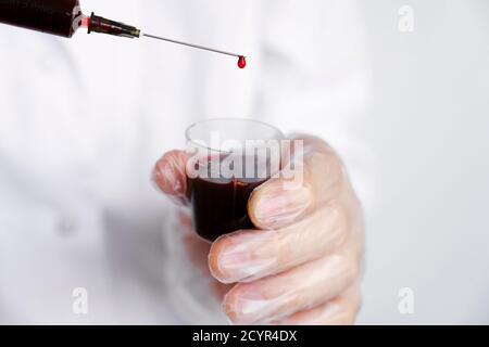 Médecin ou laboratoire scientifique dans un équipement de protection individuelle tenant une seringue remplie de sang, en se concentrant sur une goutte unique qui s'écoule de l'extrémité de l'aiguille Banque D'Images