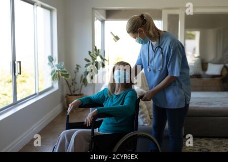 Femme caucasienne âgée à domicile visitée par une infirmière caucasienne, assise en fauteuil roulant, portant un masque facial. Soins médicaux à domicile pendant Covid 19 Coro Banque D'Images