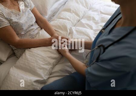 Femme caucasienne âgée à domicile visitée par une infirmière caucasienne, assise au lit, tenant les mains. Soins médicaux à domicile pendant le quar du coronavirus Covid 19 Banque D'Images