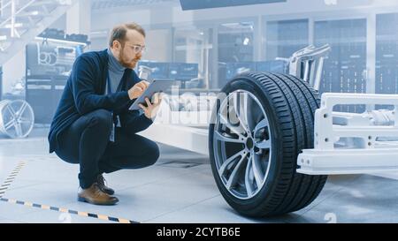 Ingénieur avec des lunettes et Beard travaille sur une tablette ordinateur à côté d'un châssis de voiture électrique prototype avec roues, batteries et moteur dans une haute technologie Banque D'Images