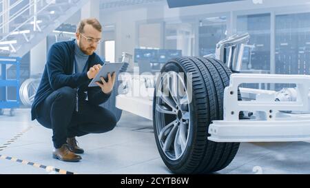 Ingénieur avec des lunettes et Beard travaille sur une tablette ordinateur à côté d'un châssis de voiture électrique prototype avec roues, batteries et moteur dans une haute technologie Banque D'Images