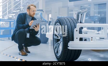 Ingénieur avec des lunettes et Beard travaille sur une tablette ordinateur à côté d'un châssis de voiture électrique prototype avec roues, batteries et moteur dans une haute technologie Banque D'Images